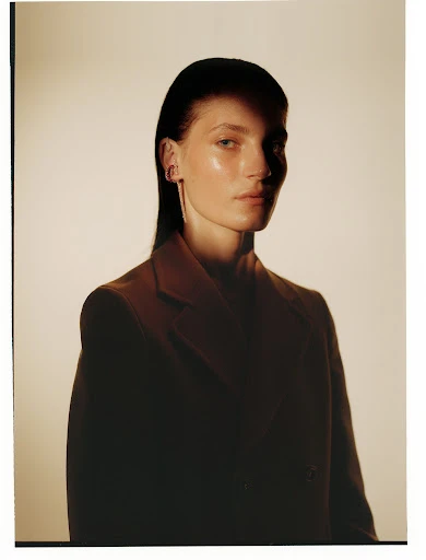 Portrait of a young adult female with dark hair, wearing an oversized brown suit jacket in a studio setting with diffused lighting.