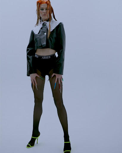 Woman with orange hair poses in black jacket, briefs, and black pantyhose against a plain backdrop in a fashion-style photograph.
