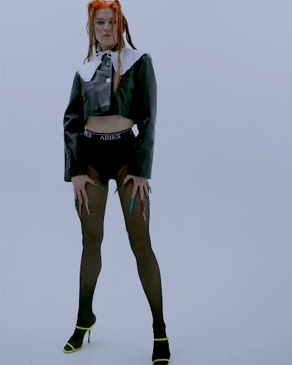 Woman with orange hair poses in black jacket, briefs, and black pantyhose against a plain backdrop in a fashion-style photograph.