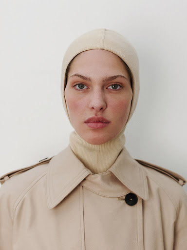 Woman in a neutral-toned trench coat and knit balaclava poses against a white background in a close-up studio portrait.