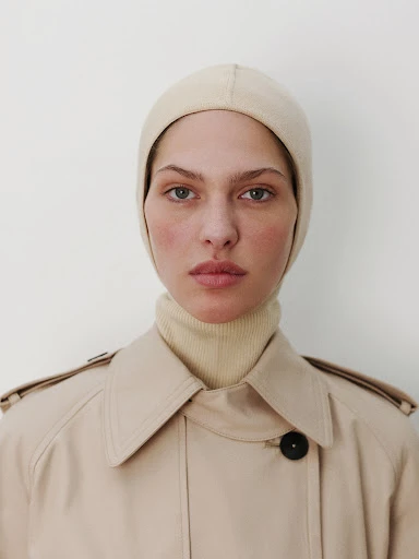 Woman in a neutral-toned trench coat and knit balaclava poses against a white background in a close-up studio portrait.