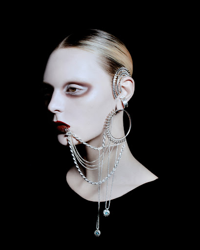 Conceptual beauty shot of a person wearing a large decorative silver earring and dark lipstick, against a stark black background.