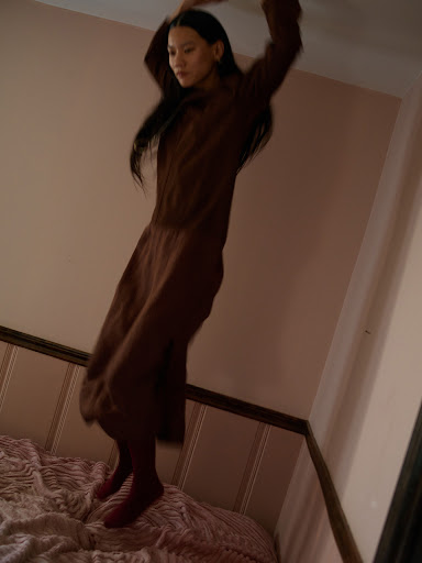 A woman with long dark hair wearing a brown dress jumping on a bed in a pink bedroom styled by Mari Siviakova