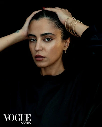 Close-up portrait of a brunette woman with hands on her head, wearing gold bracelets, against a dark background with Vogue Arabia logo.