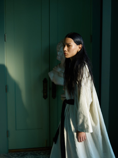 Asian woman in linen dress posing next to classical bust against muted green wall and door with soft window light and long shadow.