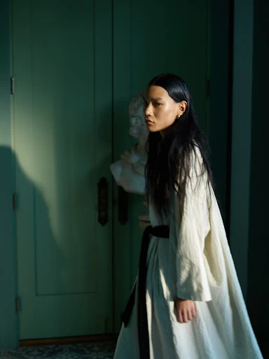 Asian woman in linen dress posing next to classical bust against muted green wall and door with soft window light and long shadow.