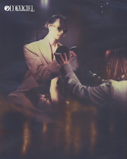 Person in blazer and sunglasses holds a mirror in a dimly lit setting with 'L'Officiel' logo visible, evoking a retro fashion vibe.