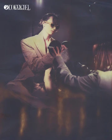 Person in blazer and sunglasses holds a mirror in a dimly lit setting with 'L'Officiel' logo visible, evoking a retro fashion vibe.