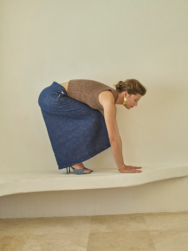 Woman in a brown knit top and denim skirt bending over a white surface with light tan walls and flooring, wearing large gold earrings.
