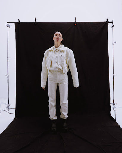 Model standing centrally in a cream denim jacket and matching pants against a dark backdrop on metal studio stands