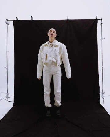 Model standing centrally in a cream denim jacket and matching pants against a dark backdrop on metal studio stands