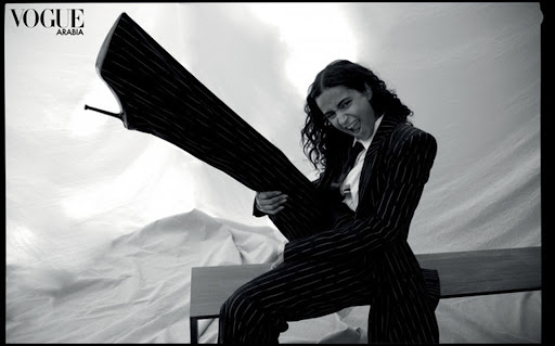 Black-and-white fashion shot of a woman in a pinstripe suit playfully holding a thigh-high boot against a textured backdrop.