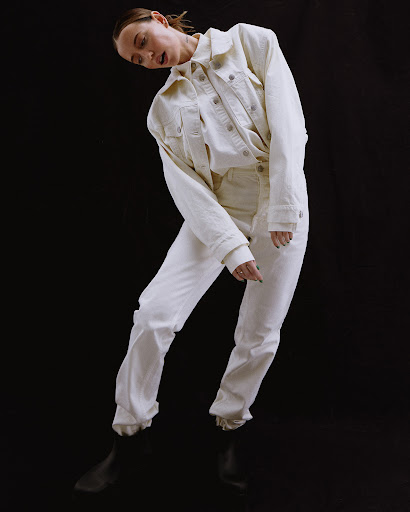 Woman in white denim jacket and pants posing against a black backdrop, fashion portrait. Stylish, confident stance and modern monochrome.