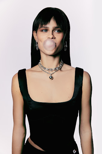 Portrait of a young woman with dark hair blowing a bubble with pink bubblegum. She is wearing jewelry and a black top in a white studio.