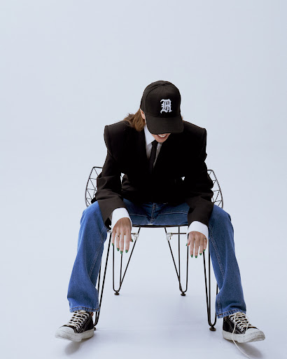Woman in jeans and black blazer sitting on a wire chair with a cap indoors against a white background in a studio setting.