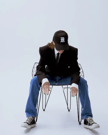 Woman in jeans and black blazer sitting on a wire chair with a cap indoors against a white background in a studio setting.