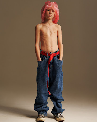 Child stands in oversized jeans and sneakers with a red belt and pink plastic bag on their head against a solid background.