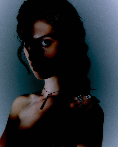 Portrait of a woman with dark hair wearing a necklace and rings, captured in a dark and moody atmosphere with a blueish background.