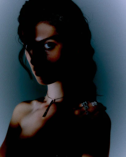 Portrait of a woman with dark hair wearing a necklace and rings, captured in a dark and moody atmosphere with a blueish background.
