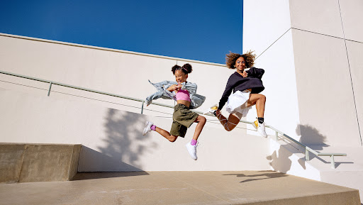 Two young women jump in the air in front of a modern white building against a bright blue sky, smiling and mid-motion.