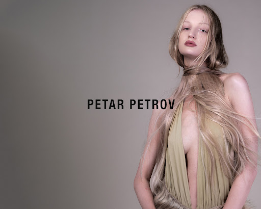 A young adult fashion model poses in a studio with long blonde hair draped around her neck and shoulders while wearing a low cut dress.