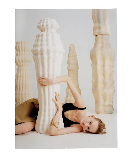 Woman in black top and brown skirt lounges against a large sculptural object with textured surface in a photo studio setting.