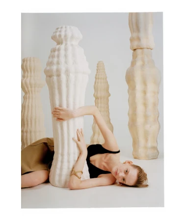 Woman in black top and brown skirt lounges against a large sculptural object with textured surface in a photo studio setting.