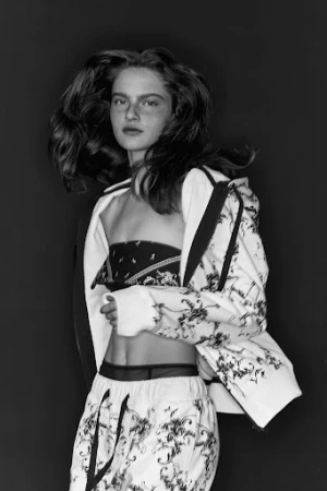 Monochrome fashion portrait of a model with voluminous dark hair wearing a printed tracksuit jacket and matching trousers against a dark background.