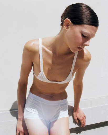 A young woman with dark hair, wearing a white bra top and white shorts, stands near a white wall, bathed in bright, neutral light.
