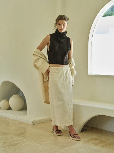 Adult woman poses indoors wearing a dark sleeveless turtleneck, cream linen skirt, and sandals, with a beige coat draped over her shoulder.