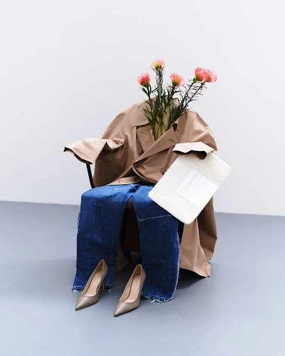 Armchair draped with a tan trench coat, blue jeans, high heels and a white package, and accented with flowers, in a white photo studio.