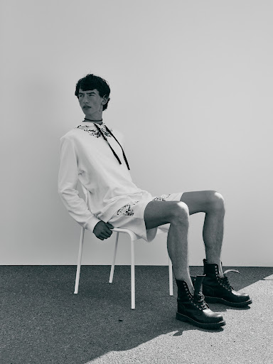 A young man sits on a white stool wearing a white hoodie and shorts with black combat boots against a plain wall backdrop.