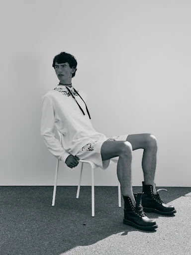 A young man sits on a white stool wearing a white hoodie and shorts with black combat boots against a plain wall backdrop.
