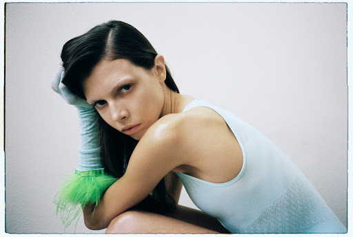 Portrait of a young woman with dark hair wearing a light blue top and gloves with green feathers, looking at the camera with a serious expression.