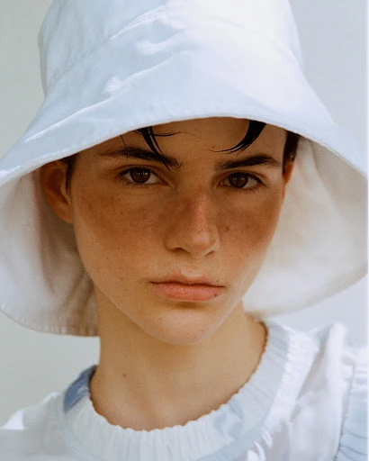 Close-up portrait of a young adult woman with freckles, wearing a white bucket hat and a white top, set against a neutral background.