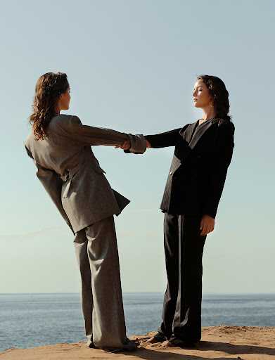Two women in suits hold hands on a cliff overlooking the ocean under a clear blue sky, showcasing fashion and connection in nature.