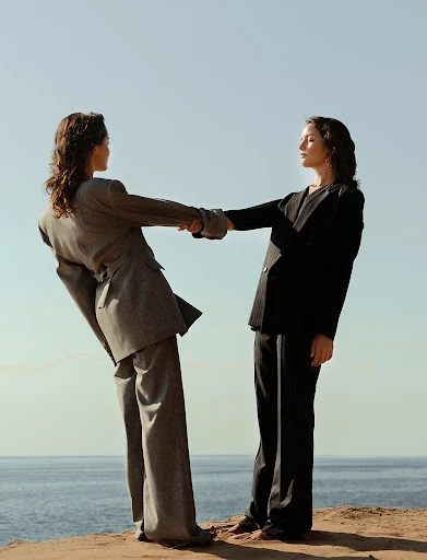 Two women in suits hold hands on a cliff overlooking the ocean under a clear blue sky, showcasing fashion and connection in nature.