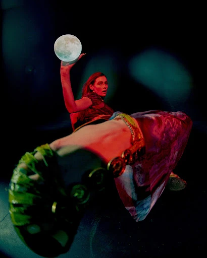 Woman in costume holds a glowing orb in a dark setting with red and blue lighting, creating a surreal and theatrical atmosphere.