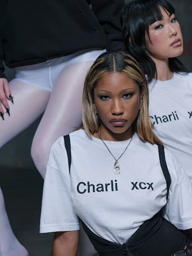 Two women pose in a studio. One woman is in focus wearing a t-shirt with 'Charli XCX' printed. Soft studio lighting.