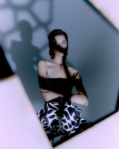 Portrait of a young woman in a black top and cow-print skirt sitting in front of a light blue wall with a geometric shadow.