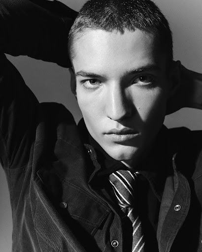 Monochrome portrait of a young adult man wearing a corduroy jacket and striped tie, gazing at the camera with his hands behind his head.