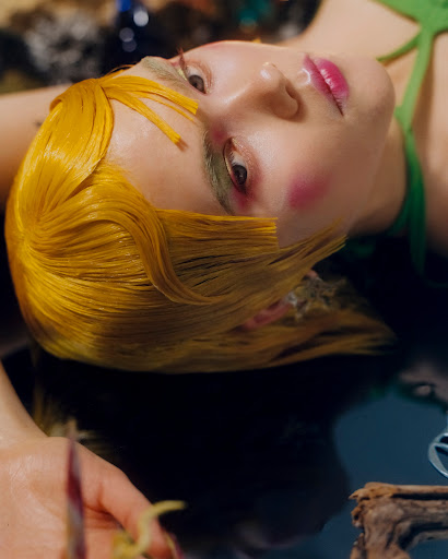 Female fashion model with yellow hair and green dress lies in a pool of water with soft lighting and artistic makeup in a studio photoshoot.