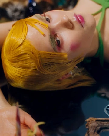 Female fashion model with yellow hair and green dress lies in a pool of water with soft lighting and artistic makeup in a studio photoshoot.