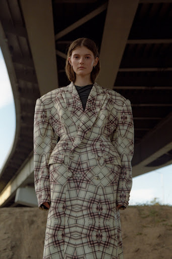 A woman stands beneath a bridge wearing a patterned skirt suit in a medium shot with low-angle framing, creating a stylish portrait.