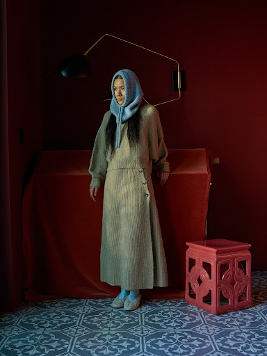 A woman wearing a beige sweater, matching long skirt, and a blue head covering, stands in a room with red walls and a patterned floor.