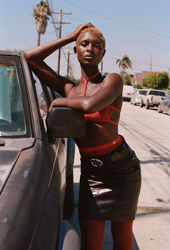 A dark-skinned woman with blonde hair wearing a red bra and black skirt leans against a car in a city street during daytime.