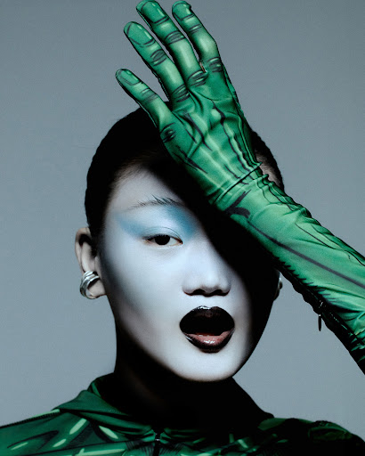 Eye-level portrait of a model with theatrical green hand and arm makeup, glossy black lips, and blue eyeshadow against a grey backdrop.