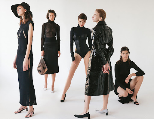 Five women in a photo studio wearing black dresses, a bodysuit, and a patent leather coat, posing on a light background for a fashion editorial.