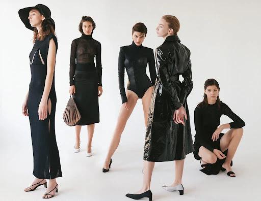 Five women in a photo studio wearing black dresses, a bodysuit, and a patent leather coat, posing on a light background for a fashion editorial.