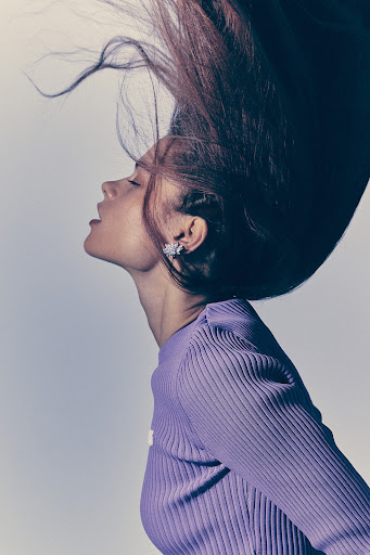 Profile portrait of a young adult woman with her head tilted back, her hair flowing upward, wearing a purple ribbed sweater and flower earrings.